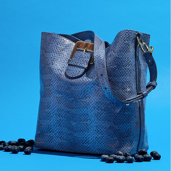 A slouchy blue python print bucket bag featuring a wide shoulder strap with a large statement buckle. The bag is positioned on a solid blue background surrounded by scattered fresh blueberries.