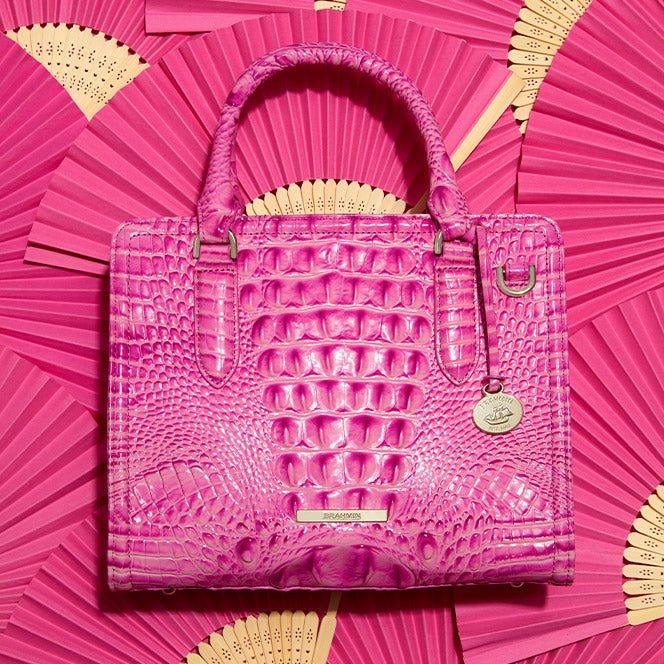 A structured bright pink handbag featuring glossy crocodile embossed leather and rolled top handles. The square satchel is displayed flat against a decorative background of matching pink folding fans.