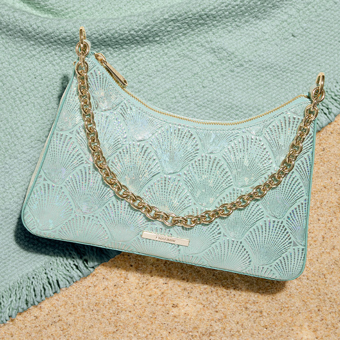A curved mint green shoulder bag with an iridescent scalloped shell pattern and a gold link chain strap. The bag rests on a patch of sand next to a soft green textured towel.