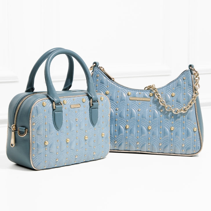 Two Brahmin handbags in soft powder-blue leather with gold studded detailing: a structured mini satchel with double handles and a matching curved shoulder bag accented with a gold chain strap, styled in front of a white wall.