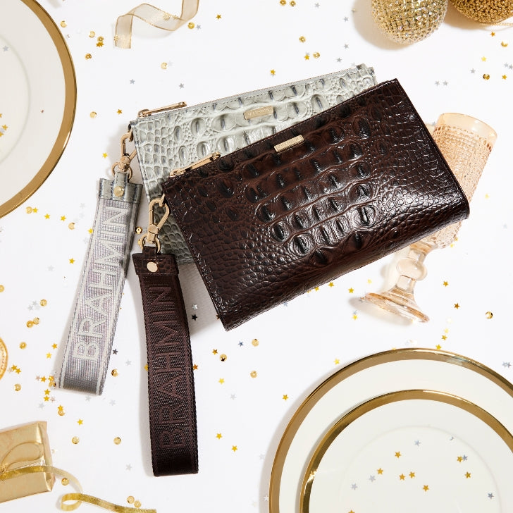 Brahmin croc-embossed wristlets in dark chocolate brown and pearl grey arranged on a festive table setting with gold-rimmed plates, sparkling glassware, ribbons, and scattered gold star confetti.