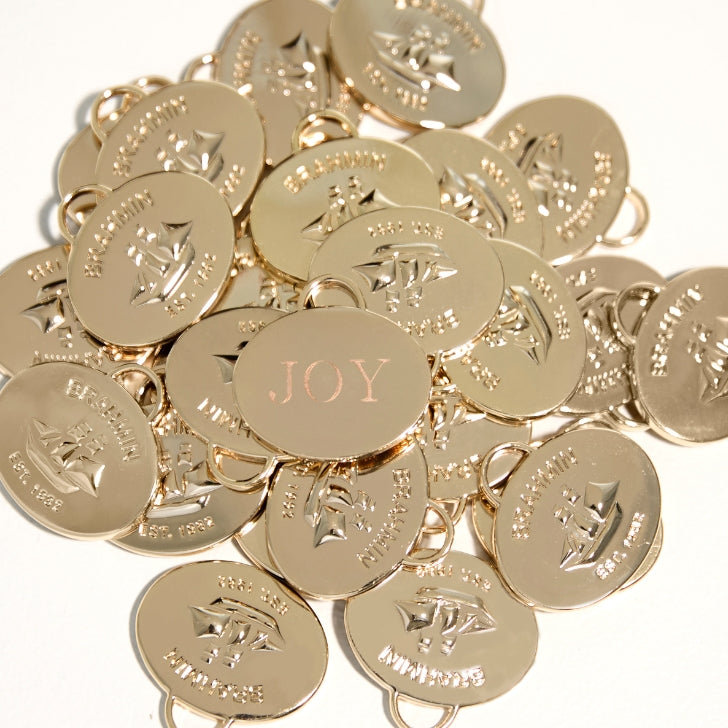 Close-up of gold Brahmin logo charms with one charm engraved with the word ‘JOY,’ reflecting a luxurious holiday detail.”