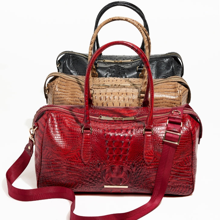 Stacked display of Brahmin croc-embossed duffle bags with a red duffle in front, taupe duffle in the middle, and black duffle behind, all featuring structured handles and gold hardware.