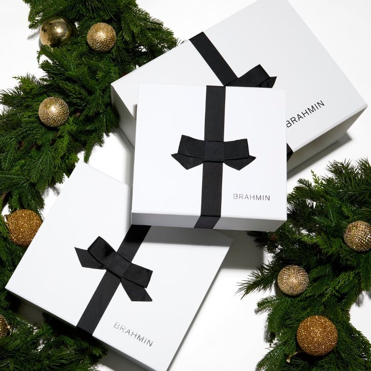 Overhead view of several stacked white Brahmin gift boxes tied with elegant black satin bows and surrounded by festive Christmas tree branches and gold ornaments.