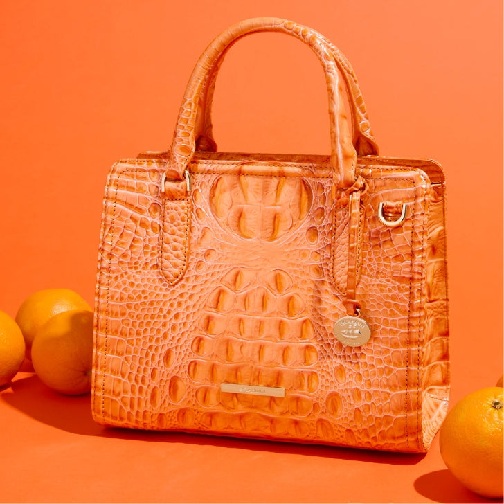 Orange croc-embossed leather satchel handbag with a structured top-handle design, raised scale detailing, gold-tone hardware, and detachable shoulder strap, styled with fresh oranges on a vibrant backdrop