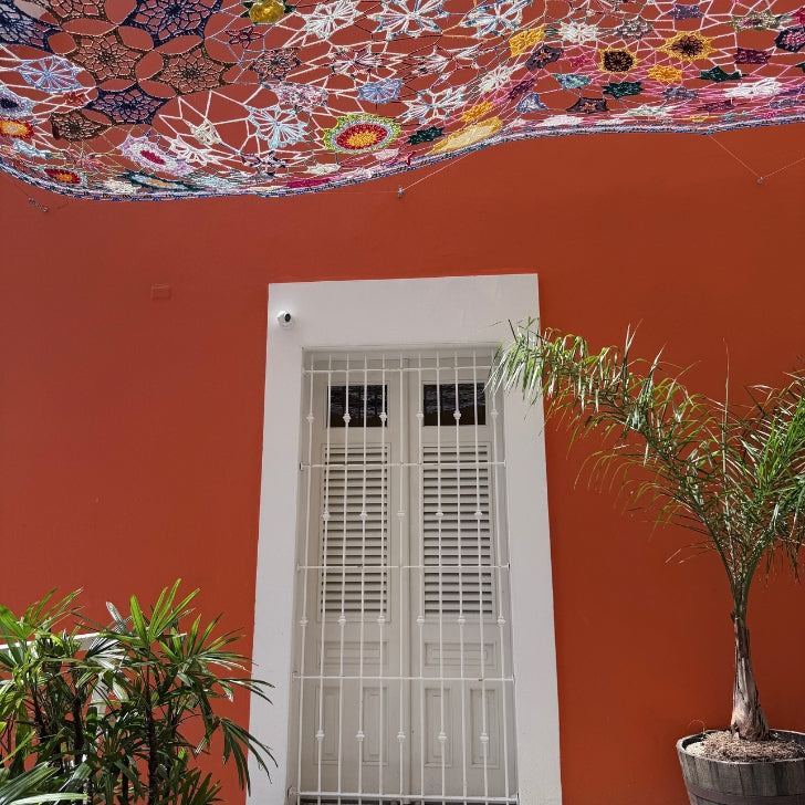 Sunlit courtyard with a white shuttered door framed by terracotta walls, tropical potted plants, and a vibrant multicolored lace canopy casting patterned shadows overhead.