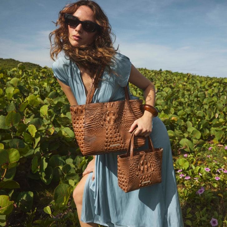 Brown crocodile-embossed leather tote with a matching mini crossbody bag, showcasing rich texture and structured design, styled outdoors with a flowing blue dress against a lush green landscape.