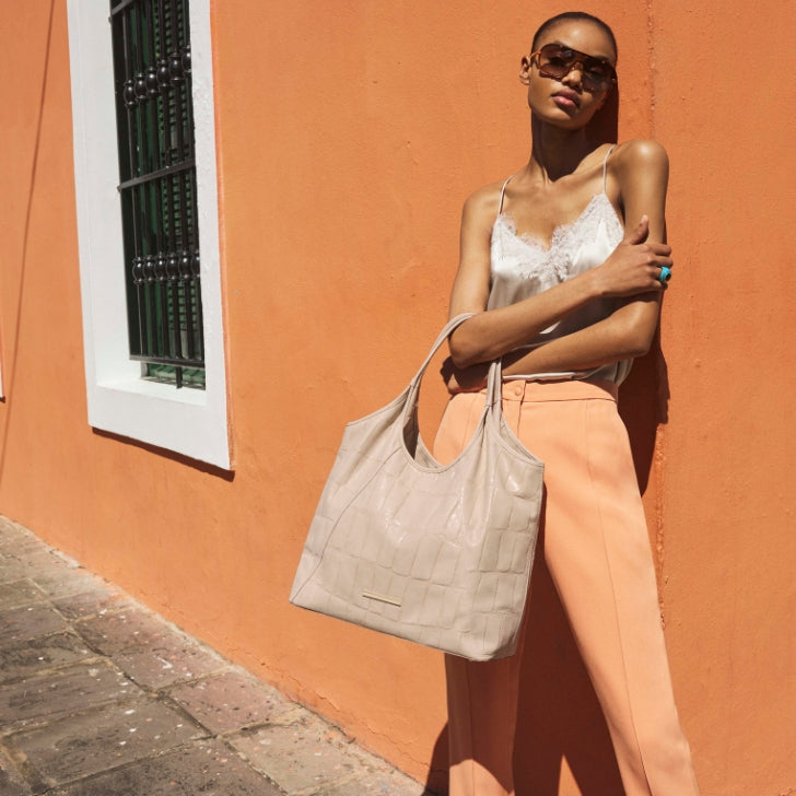 Neutral taupe crocodile-embossed leather tote bag with double top handles and a spacious interior, styled with tailored peach trousers and a silk camisole against a warm terracotta wall.