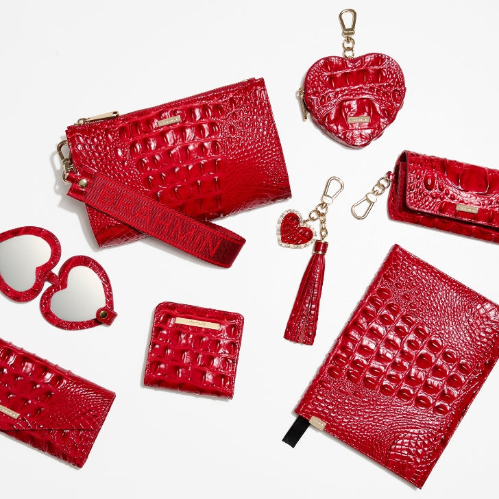 Red croc-embossed leather accessory assortment including zip wristlet with logo strap, compact wallet, coin pouch, heart-shaped mirror, heart charm keychain, and leather tassel, arranged on a white background
