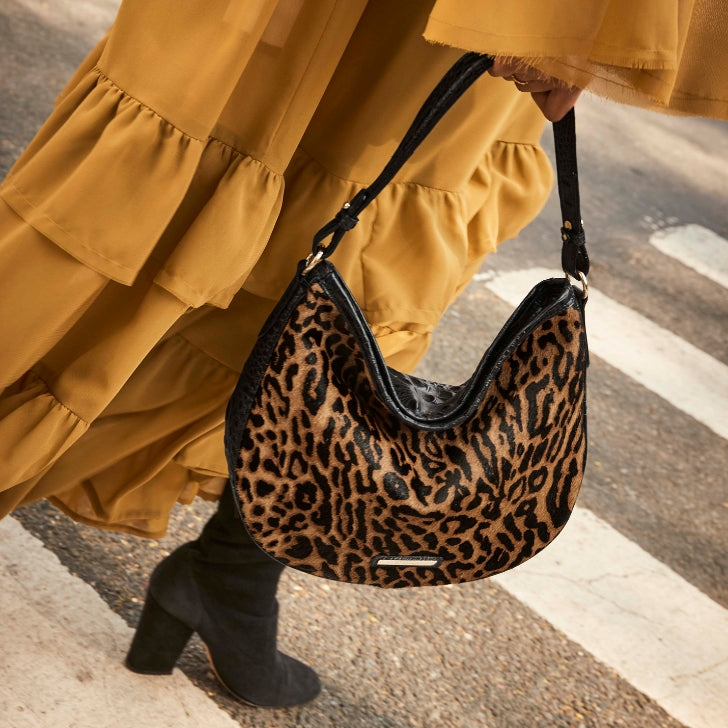 Close-up of a Brahmin leopard-print shoulder bag with black trim carried by a woman in a flowing mustard dress while walking across a city street.