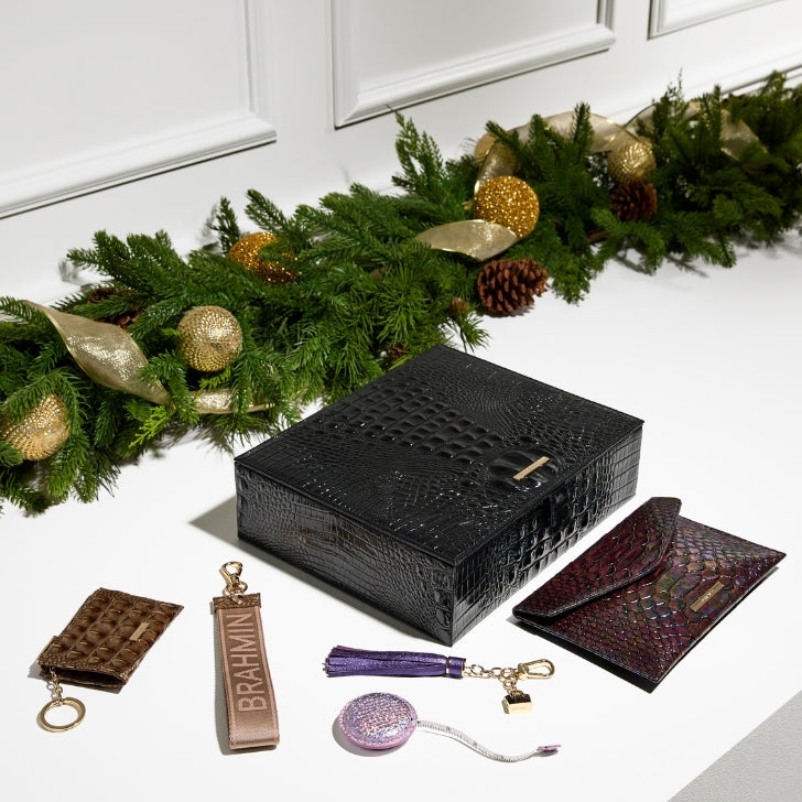 Brahmin black croc-embossed gift box displayed with assorted small leather goods—including a brown card case, pink glitter coin pouch, purple tassel keychain, gold lock charm, branded strap, and a multicolor envelope wallet—arranged in front of a holiday garland with gold ornaments and pine cones.
