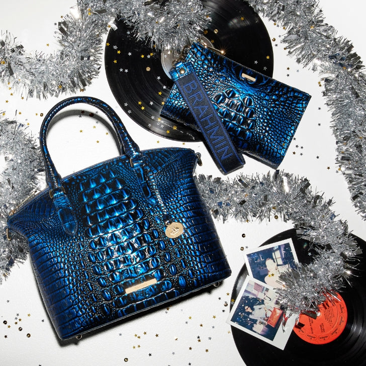 Brahmin sapphire blue croc-embossed satchel paired with a matching blue wristlet, styled on a festive white background with vinyl records, silver tinsel, and scattered gold star confetti.