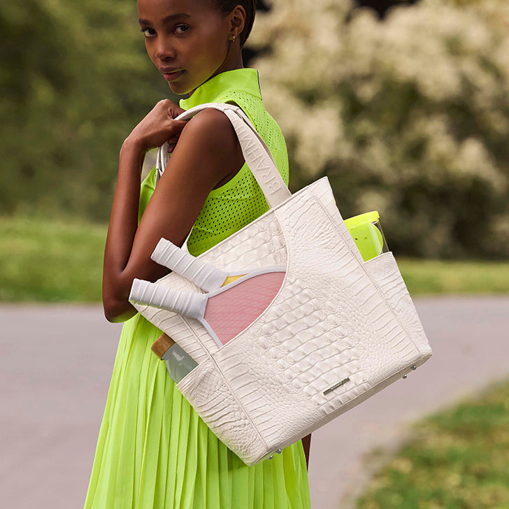 Woman carrying a Brahmin Mollie Tote in textured white leather, styled with a neon green dress outdoors. The spacious tote features exterior pockets holding yoga essentials, a water bottle, and accessories.