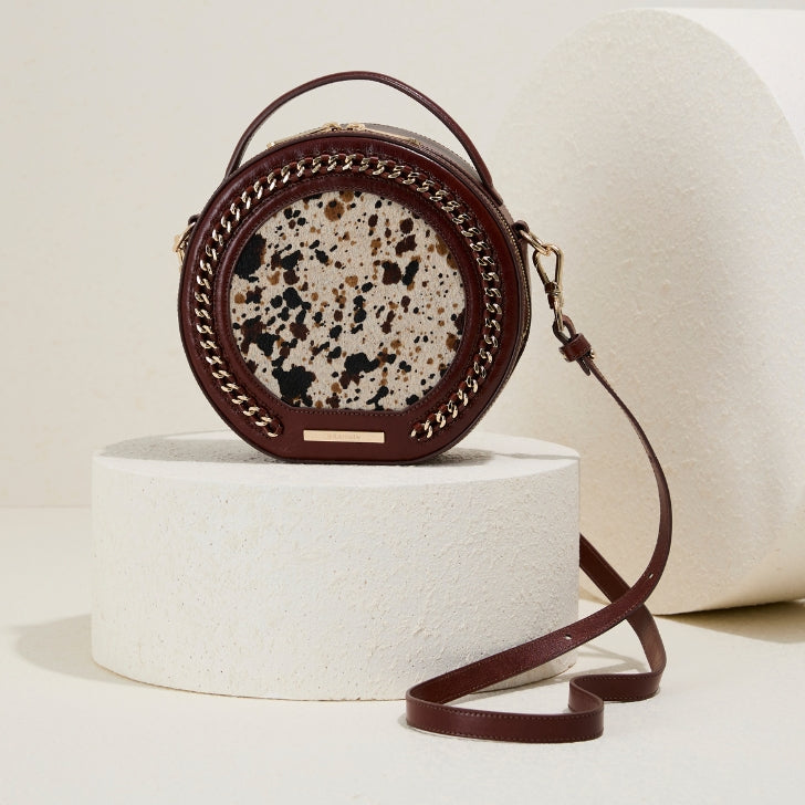 Brahmin Cognac Caspian Lane round leather crossbody bag with speckled tan and black pony hair front panel, gold-tone chain trim and adjustable strap, displayed on a white pedestal.