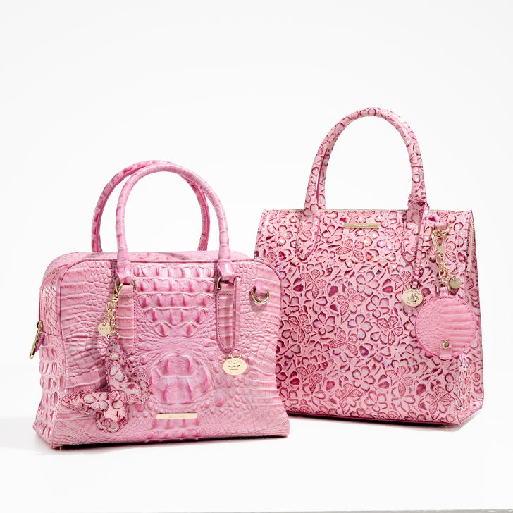 Side-by-side Brahmin Fearless Collection handbags: Caroline tote in pink floral embossed leather and Marissa satchel in pink croc-embossed leather, both with gold hardware and signature charms, limited-edition designs created to support the National Breast Cancer Foundation.