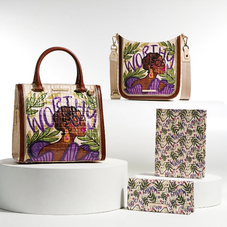 Brahmin's Lilac Worthy collection displayed on white platforms, featuring a handbag, crossbody bag, wallet, and journal with bold purple tones and an illustrated affirmation design.