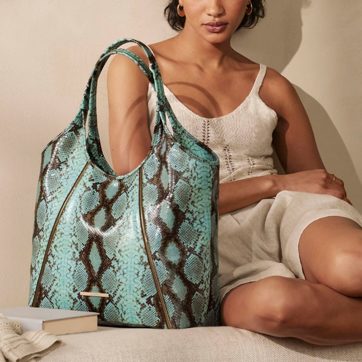 Brahmin Iona slouch tote in turquoise python-embossed leather with draped double handles, showcased next to a neutral linen sofa.