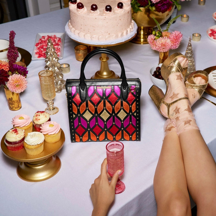 A bold geometric tote in metallic pink, orange, and gold leather sits on a holiday dessert table with cupcakes, flowers, and a frosted cake, styled with golden platform heels and star-patterned sheer socks for a playful party vibe.