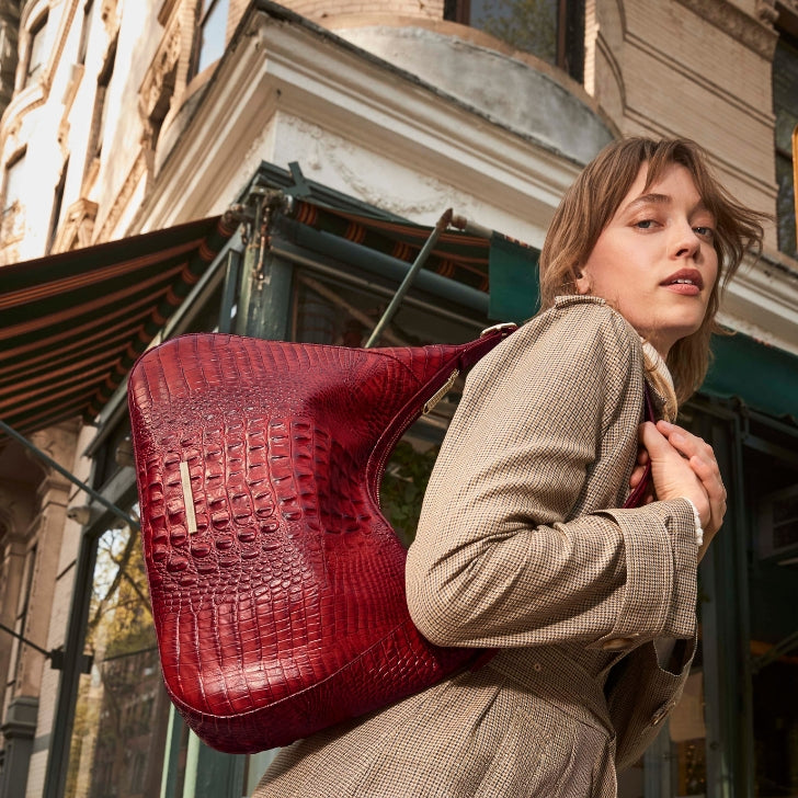Woman in a tan blazer carrying a large glossy burgundy croc‑embossed leather hobo bag over her shoulder on a city sidewalk.