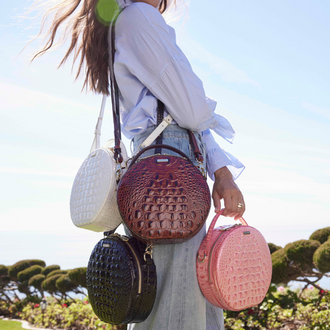 A woman wearing a light blue blouse and jeans carrying multiple Brahmin Lane crossbody bags in different colors, including white, brown, black, and pink, with a scenic outdoor background.
