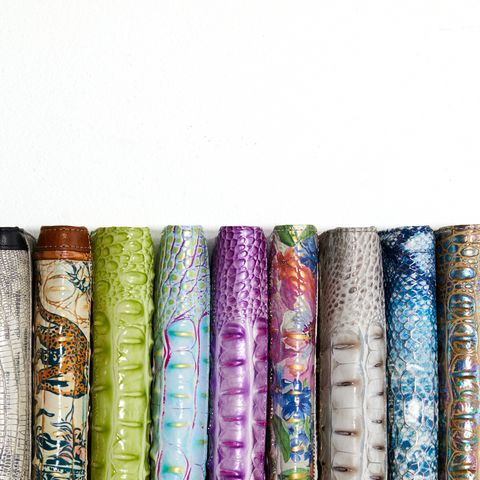 A row of Brahmin Leather long wallets featuring various colorful and textured designs, including patterns resembling scales, leather, and abstract motifs in a range of colors from metallic and neutral tones to vibrant greens, purples, and blues.